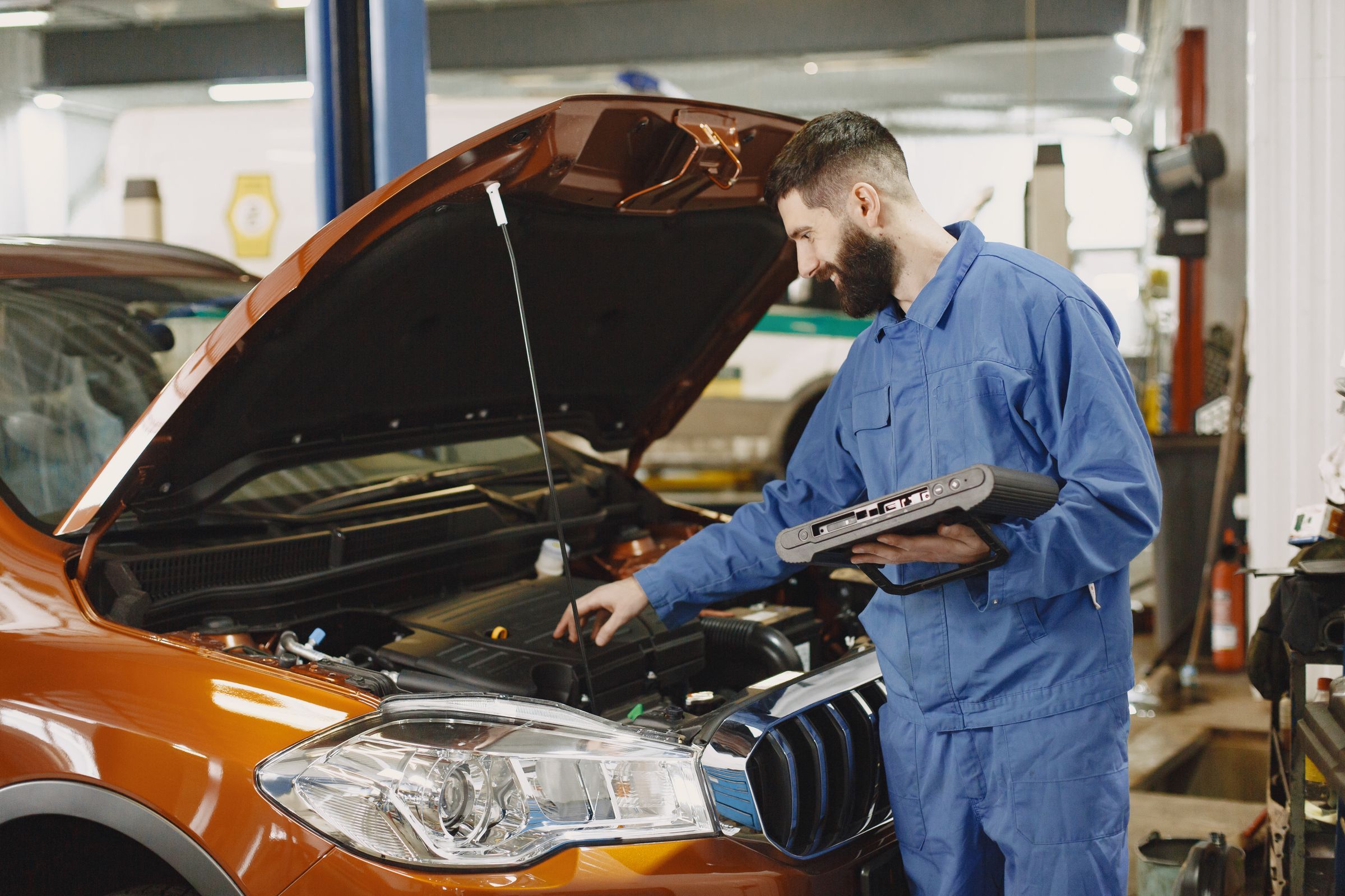 A smiling technician using a diagnostic tablet beside an open hood in an auto repair shop